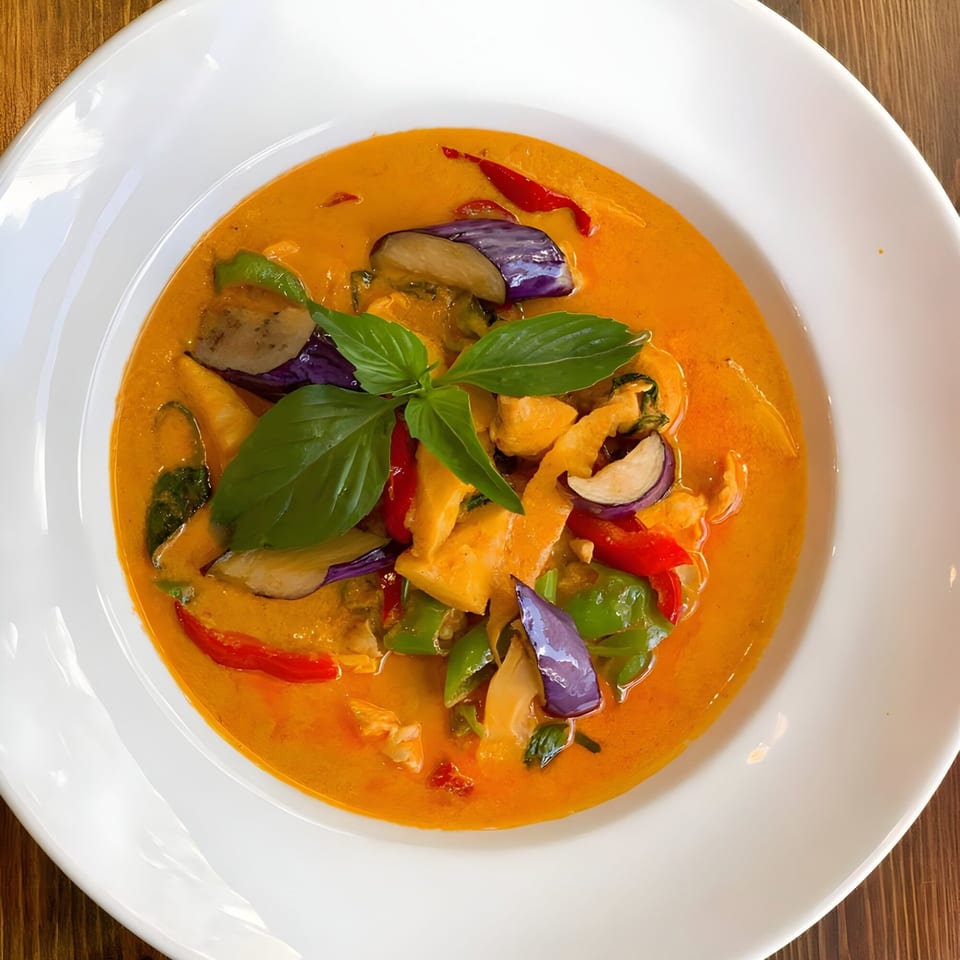One More Thai red curry with chicken, eggplant, bell peppers, and basil in a white bowl.