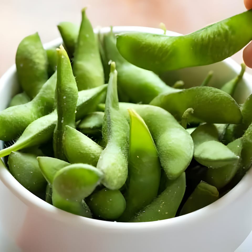 Close-up of a white bowl filled with green edamame pods, one being picked up.